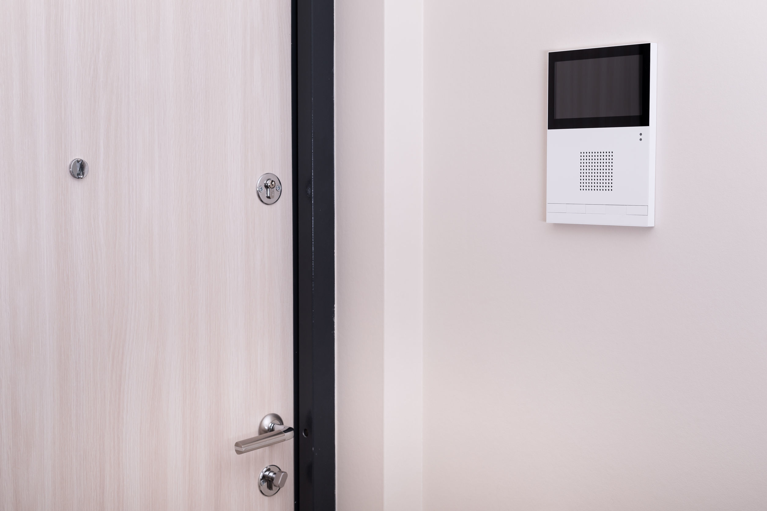 Moderne Türsprechanlage an einer hellen Wand neben einer Holztür mit Schloss und Griff – Teil der Sicherheitslösungen von Limburger Sicherheitstechnik für Zutrittskontrolle und Einbruchschutz.
