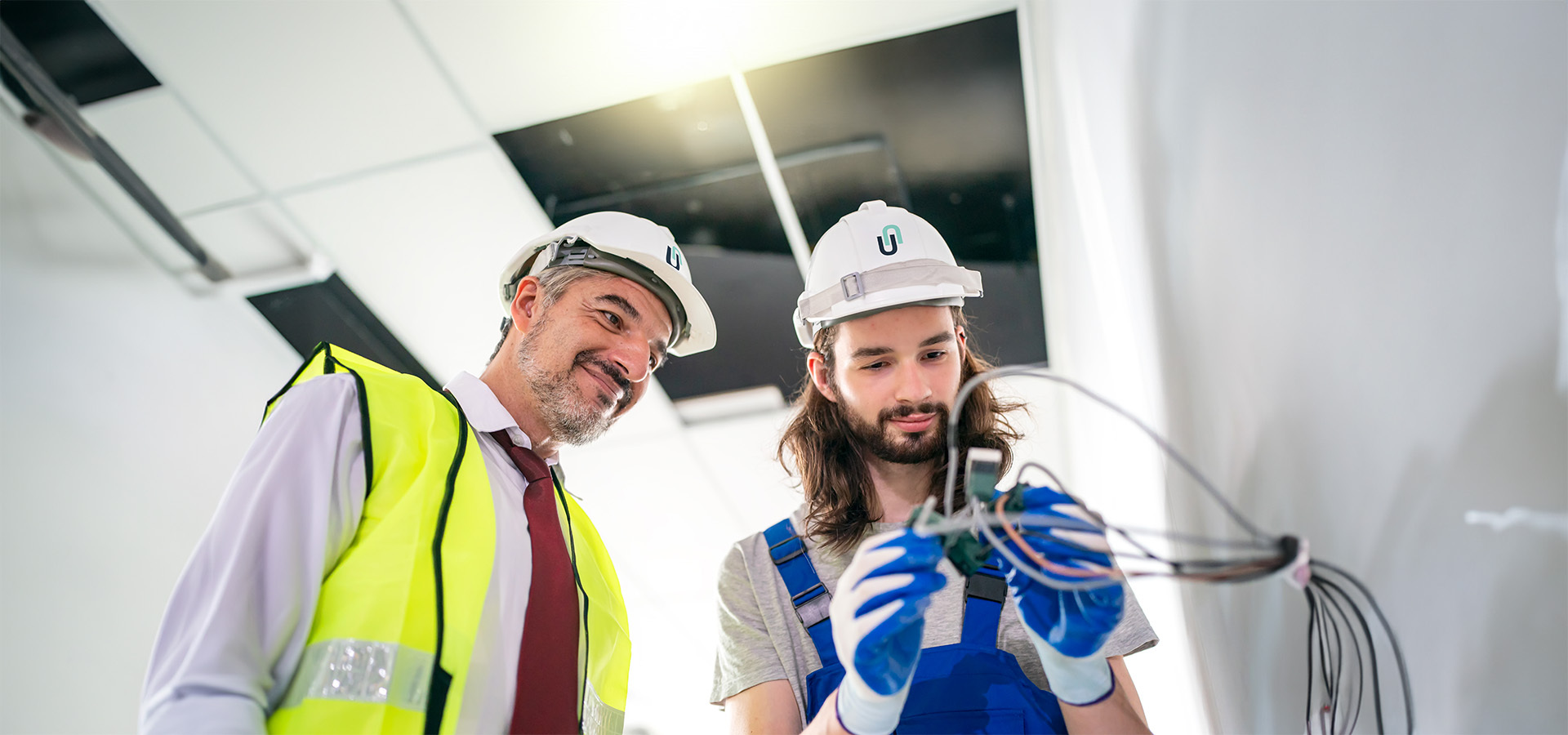 Ein erfahrener Elektroinstallateur in Warnweste und sein Azubi in Arbeitskleidung arbeiten an Elektrokabeln in einem Büro – symbolisch für die Sicherheits- und Elektrotechnikleistungen von Limburger Sicherheitstechnik.