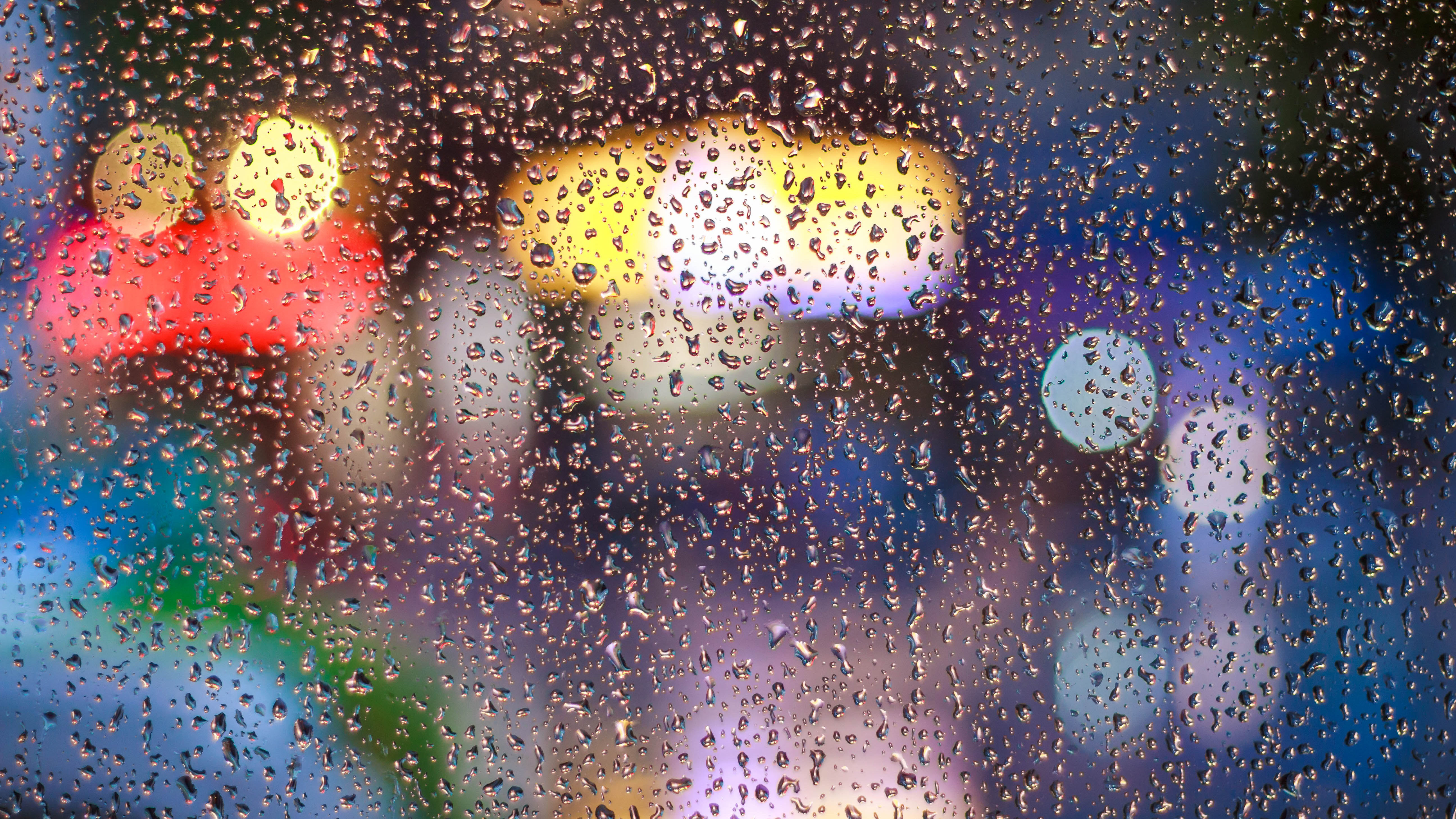 Regentropfen auf einem Fenster mit verschwommenen bunten Lichtern im Hintergrund, symbolisierend den Schutz vor Einbruch in regnerischen Nächten.