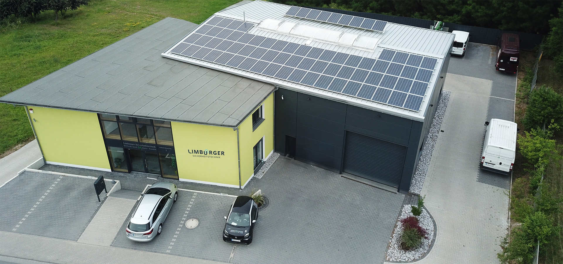 Luftaufnahme eines modernen Bürogebäudes von Limburger Sicherheitstechnik mit Solarpanelen auf dem Dach, Parkplatz mit Fahrzeugen und Firmenschild.