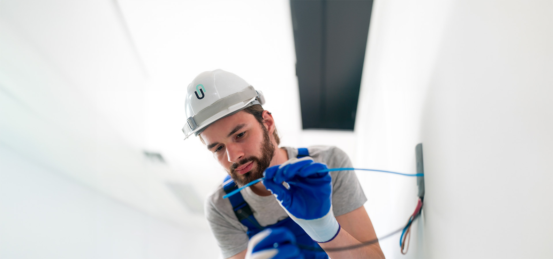 Azubi-Elektroinstallateur mit Schutzhelm und blauen Handschuhen führt Elektroarbeiten an Kabeln durch – typisch für Sicherheits- und Elektrotechnik-Dienstleistungen.