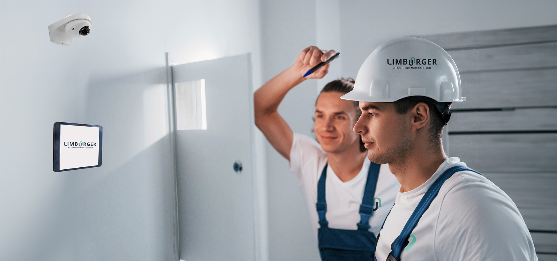 Zwei Mitarbeiter von Limburger Sicherheitstechnik arbeiten an der Installation oder Überprüfung von Sicherheitsausrüstung, darunter einer Überwachungskamera und einem Display mit dem Firmenlogo, in einem Innenraum.