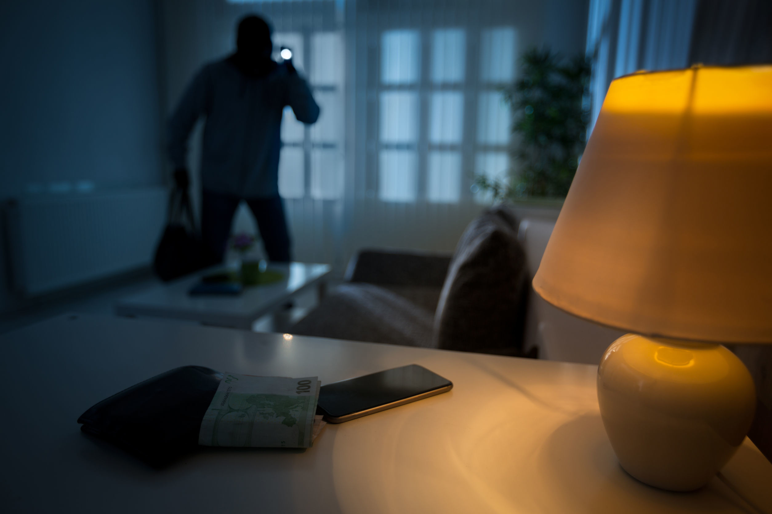 Ein Einbrecher in einem Wohnzimmer mit Geldbörse, Smartphone und Lampe auf dem Tisch – ein Beispiel für die Notwendigkeit von professionellem Einbruchschutz.