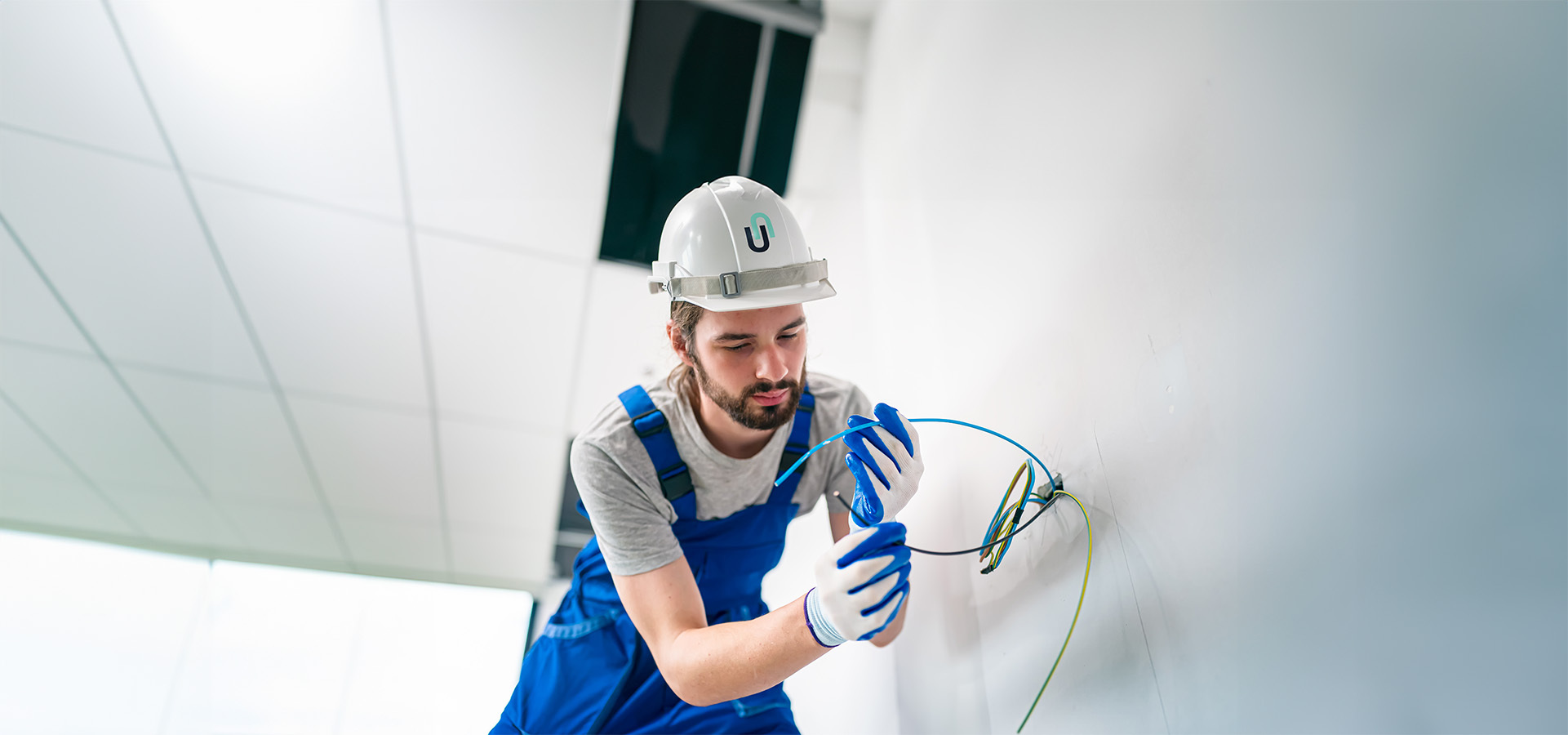 Ein Elektroinstallateur-Azubi in blauer Arbeitskleidung und weißem Helm arbeitet an der Kabelinstallation an einer Wand, typisch für Sicherheits- oder Sicherheitstechnikprojekte.