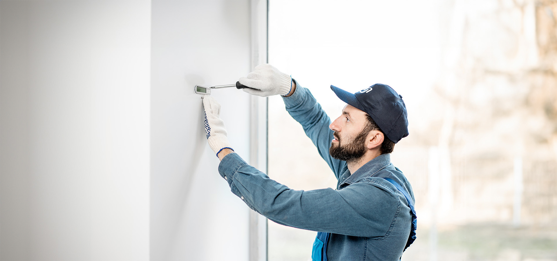 Ein Sicherheitstechniker von Limburger Sicherheitstechnik arbeitet an der Installation einer Sicherheitskomponente an einer Wand, wobei er Handschuhe und eine Arbeitskappe trägt.