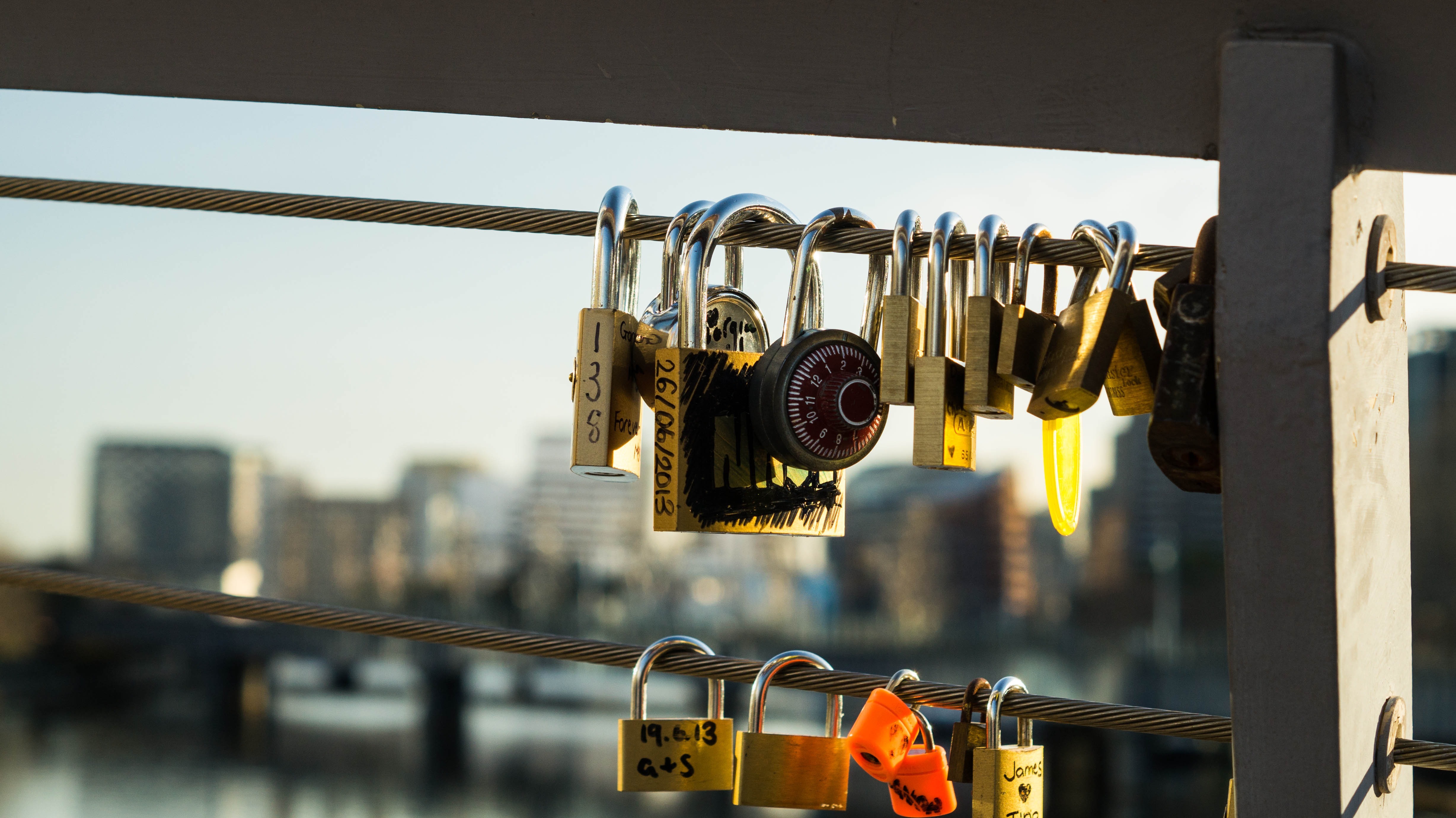 Liebesschlösser in Gold, Silber und Orange hängen an einem Brückenseil, mit einer urbanen Skyline im Hintergrund. Die Schlösser symbolisieren Liebe und Sicherheit, passend zum Thema Sicherheitstechnik.